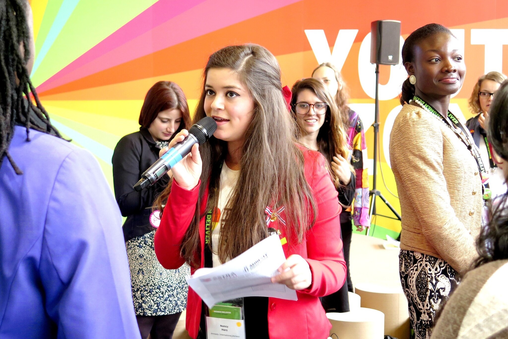  A young woman wearing a red jacket speaks into a microphone while holding notes, standing in front of a colourful rainbow-striped backdrop. Several other young people stand behind her, listening and smiling, suggesting a youth-led event or conference focused on activism or community engagement.